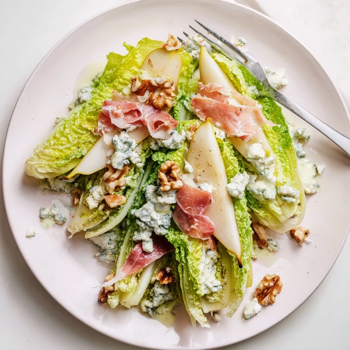 Frisée, Pear, Blue Cheese, and Crispy Prosciutto Salad is arranged elegantly on a white platter, showcasing bright greens and golden-brown crisps.  